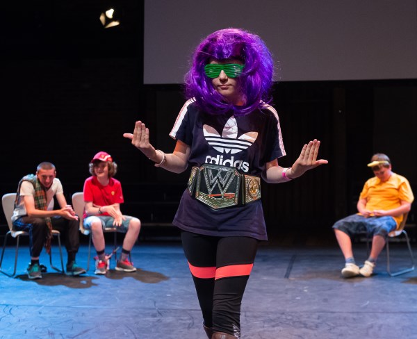 Young performers onstage, the foremost of which is wearing a purple wig and green shutter-glasses while they gesture to come closer