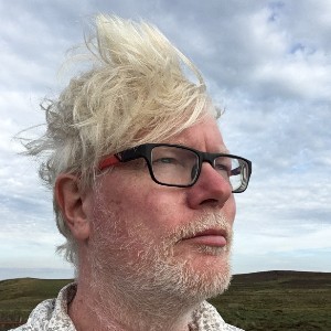 Aidan Moesby wearing glasses standing against against a blue sky with wind blowing his hair