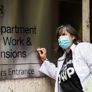 Dolly sens in doctor coat and mask putting a thermometer against the sign of the Department of Work and Pensions