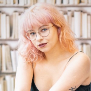 Photographer Natalie Bedkowska photographed in front of bookshelves. She wear glasses and has pink hair.