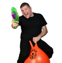 A man in a back t-shirt and trousers holds a green toy gun and bounces on an orange space hopper against a white bakground