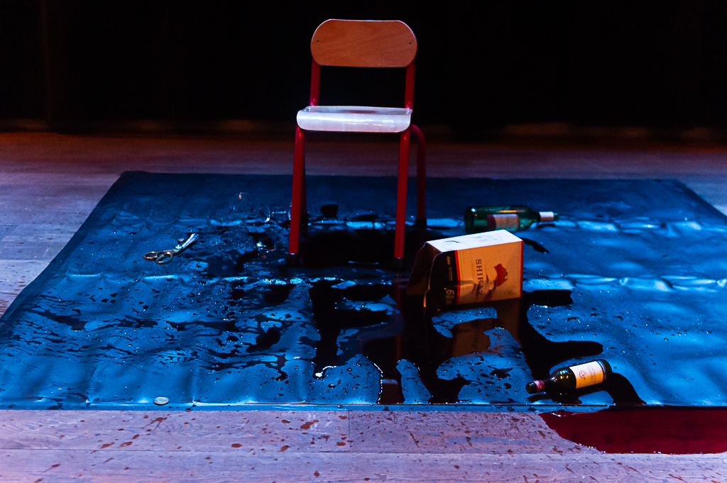 A chair on a tarpaulin splattered with wine, with some of the wine leaking off the edge onto a wood panelled floor. 