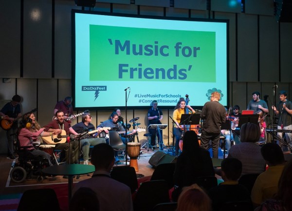 Young musicians and a conductor onstage in front of a projector screen on which it reads 'Music for Friends'