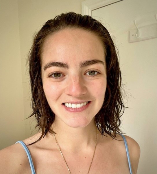 Smiling young white woman with shoulder length brown hair, green eyes and big eyebrows, wearing a light blue strappy top and gold necklace