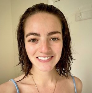 Smiling young white woman with shoulder length brown hair, green eyes and big eyebrows, wearing a light blue strappy top and gold necklace