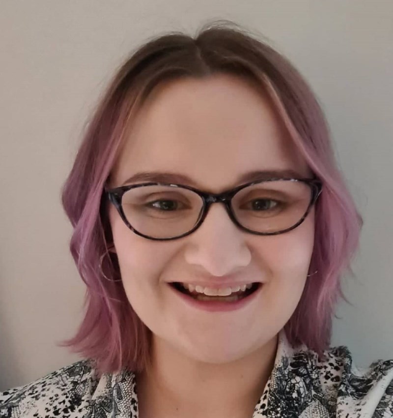 a white woman standing in front of a white wall. She has chin length hair that is dyed a lavender shade with a dark root. She is wearing glasses