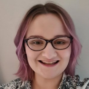 a white woman standing in front of a white wall. She has chin length hair that is dyed a lavender shade with a dark root. She is wearing glasse