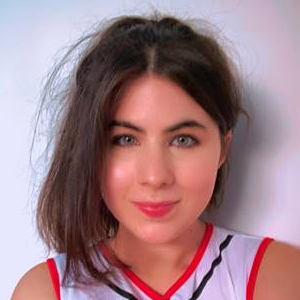 a white woman with green eyes and her straight brown hair tied up in a messy ponytail. She is wearing a cheerleader outfit.