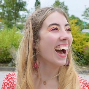 A young person in a leafy, outdoor setting with long blondish hair and brown eyes, wearing a  red dress and red flower earrings, smiling.