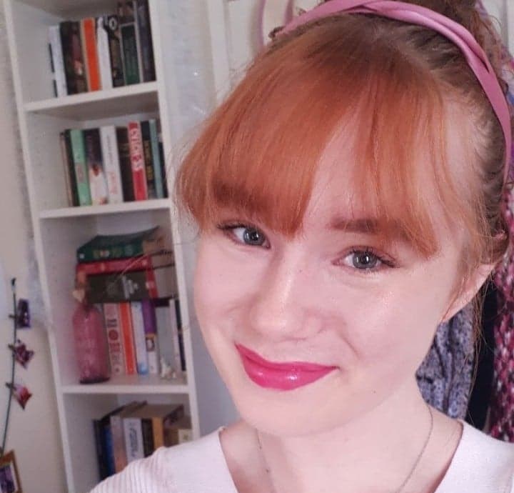 a white woman with red hair in a fringe, with bright pink lipstick on and a pink ribbon in her hair. She is smiling