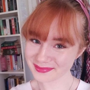 a white woman with red hair in a fringe, with bright pink lipstick on and a pink ribbon in her hair. She is smiling