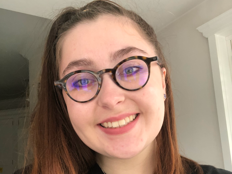 A white woman with long auburn hair in a ponytail, wearing brown-rimmed glasses, purple eyeliner, purple earrings and a black tshirt. and smiling