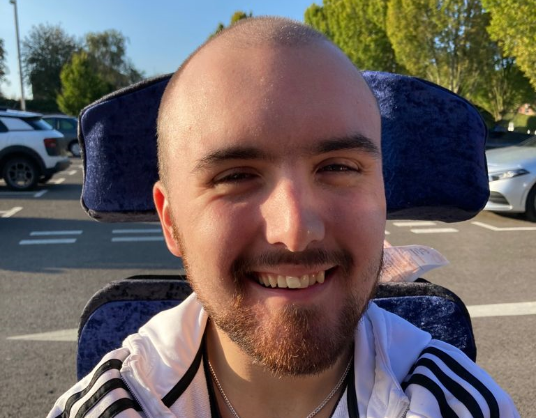 Jack is a white male in his early 20’s with a shaved head and dark brown facial hair. He wears a white tracksuit stop, silver chain and is smiling. 