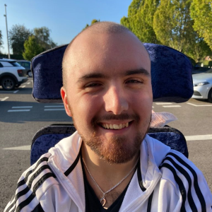 Jack is a white male in his early 20’s with a shaved head and dark brown facial hair. He wears a white tracksuit stop, silver chain and is smiling. 