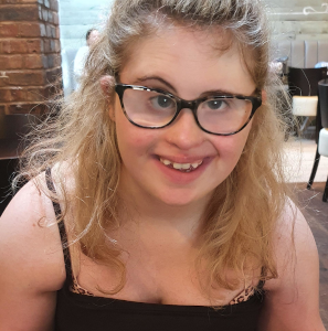 A smiling young adult wearing glasses and a vest top sat behind a plate of fish, chips and beans. She has long blonde hair