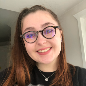 A white woman with long auburn hair in a ponytail, wearing brown-rimmed glasses, purple eyeliner, purple earrings and a black tshirt. and smiling