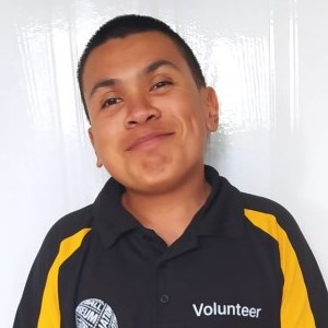 A mixed race East Asian and Bengali man with black hair wearing a black top that says 'Volunteer' alongside a logo for a Football museum