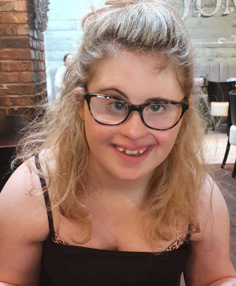 A smiling young adult wearing glasses and a vest top sat behind a plate of fish, chips and beans. She has long blonde hair