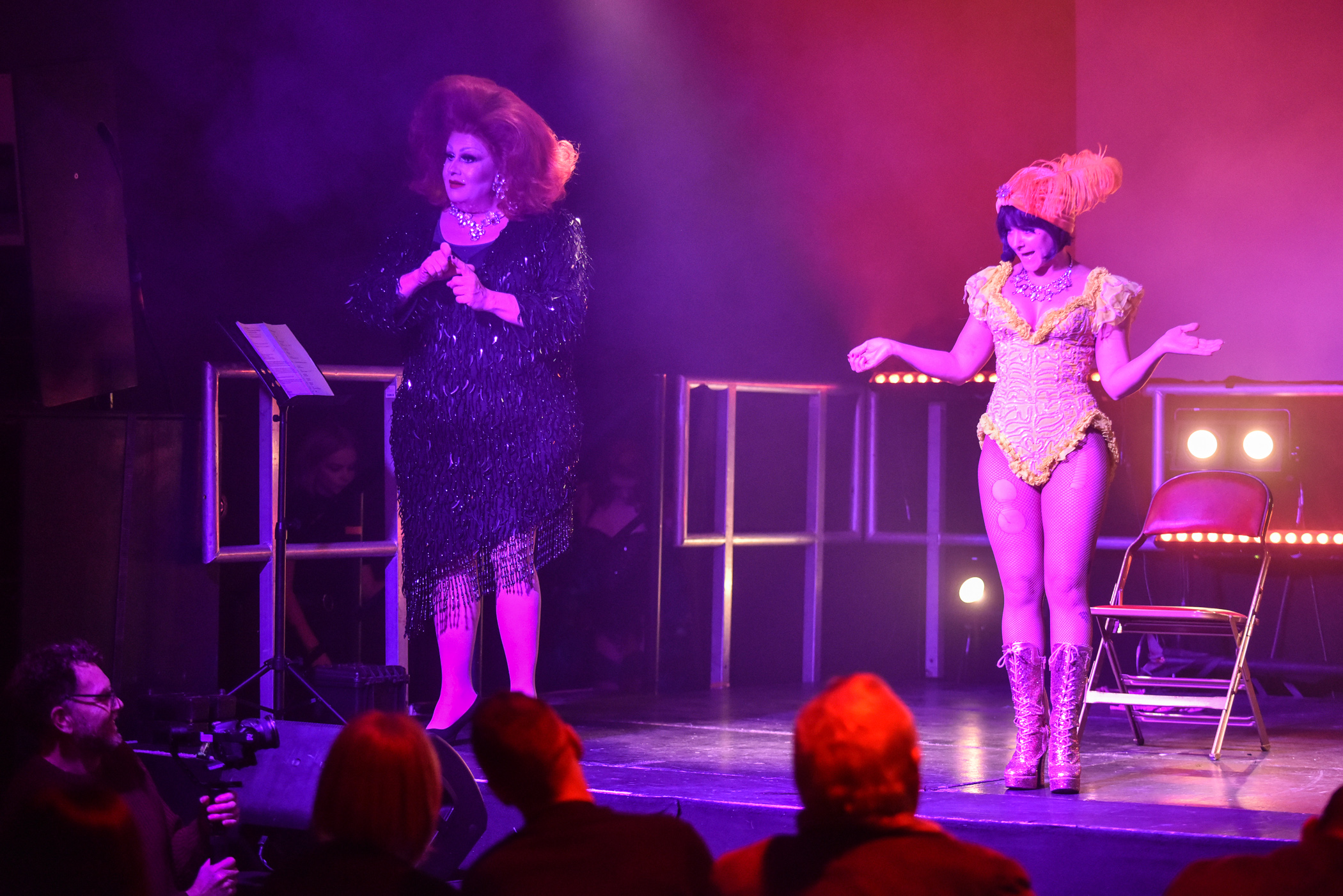 Two people onstage dressed glamorously, one as a showgirl and the other in a blue sequined-dress and large red-haired wig. 