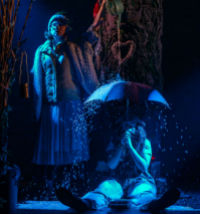 A photo of a man sitting on a dimly lit stage holding an umbrella over his head whilst a woman pours water on it from a watering can.