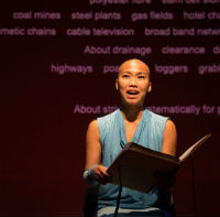 A photo of a woman on stage in a blue dress seen from the waste up reading from a large book. Behind her text is projected onto a red wall.