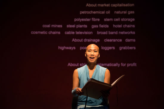 A photo of a woman on stage in a blue dress seen from the waste up reading from a large book. Behind her text is projected onto a red wall.