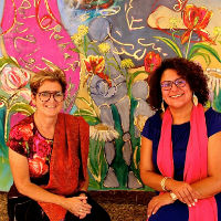 A colourful painting. In front of it sits artist Rachel Gadsden and ethnographer Dr. Yafa Shanneik