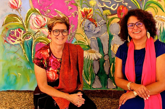 A colourful painting. In front of it sits artist Rachel Gadsden and ethnographer Dr. Yafa Shanneik