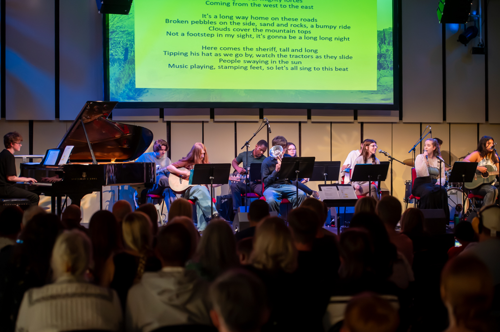 A group of young musicians are gathered on a stage. One is sat at a grand piano. Others have guitars, violin, trumpet and microphones. Behind is a yellow screen with some lyrics on it