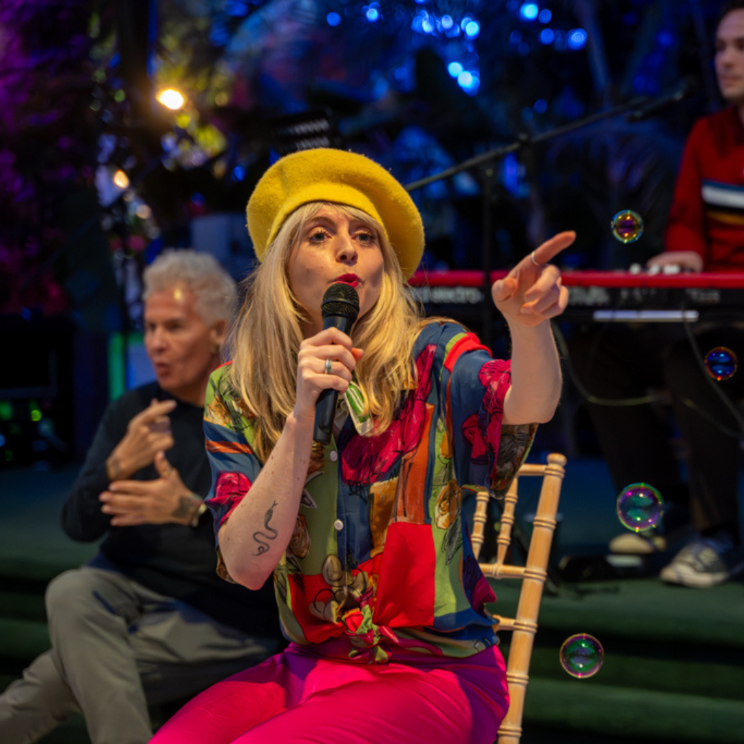 Dora is seated at Sefton Park Palm House singing into a mic pointing at a bubble floating in the air