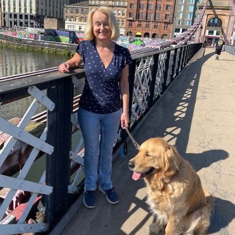 Gill is a blond woman wearing jeans and a blue top.. She stands on a bridge on a sunny day with her guide dog