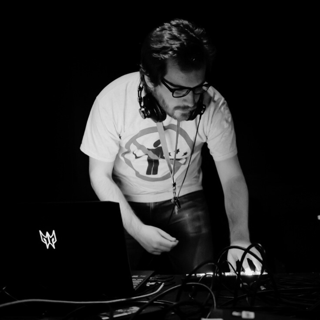 Artist Joe Strickland wearing t-shirt, jeans and glasses has dark hair and a beard.  Joe has headphones around their neck and is leaning over a table with a laptop on and lots of wires working as a digital producer