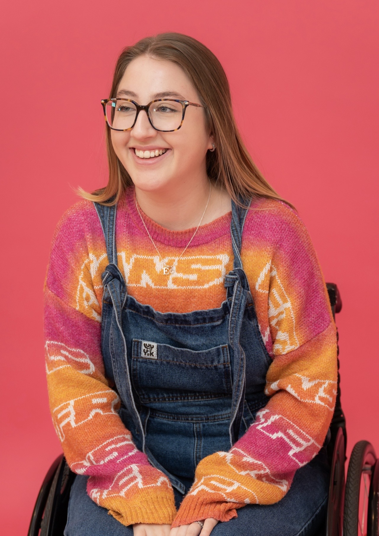 Maisy is seated in a wheelchair against a red background. She has long brown hair and glasses. She is wearing denim dungarees with colourful jumper