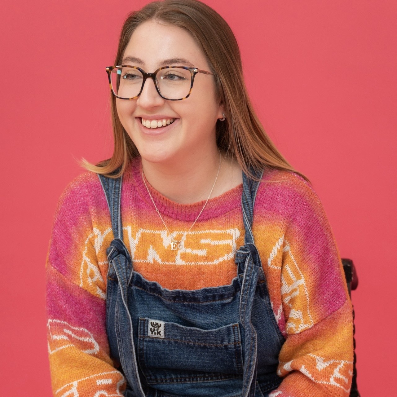 Maisy has long brown hair and glasses. She is wearing denim dungarees with colourful jumper