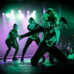 Three dancers onstage under purple and green lighting. The foremost is on their knees with their arms spread out dramatically.