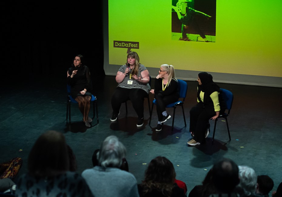 Four women onstage during a discussion. One holds a microphone and is speaking, two are listening to her and the other is a sign language interpreter