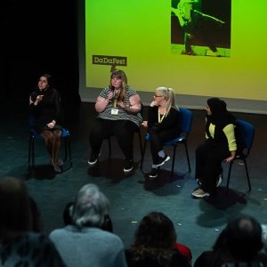 Four women onstage during a discussion. One holds a microphone and is speaking, two are listening to her and the other is a sign language interpreter