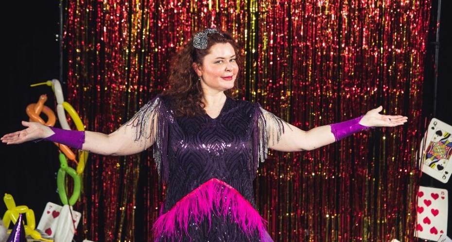 Elaine Collins, a white woman with red hair wearing a sparkly, purple fringed dress stood in front of  red and yellow sparkly fringed curtain. 