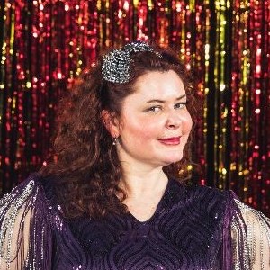 Elaine Collins, a white woman with red hair wearing a sparkly, purple fringed dress stood in front of  red and yellow sparkly fringed curtain. 