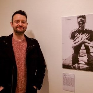 Brown haired man stood alongside a black and white photographic self-portrait showing different images of himself superimposed on one another
