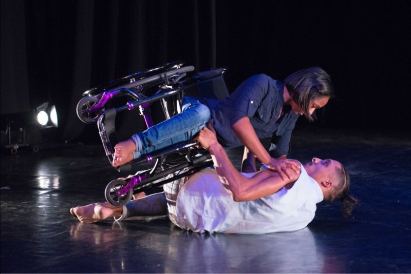 Dancers in a dark performance space. One lies on the ground, supporting the other who is a wheelchair user, as she tilts herself over him