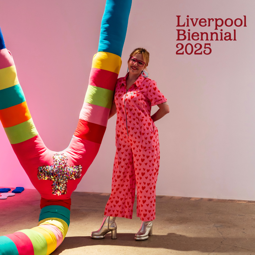Amy Claire Mills wears glasses and stands dressed in a pink jumpsuit next to a large colourful U shaped artwork covered in fabric