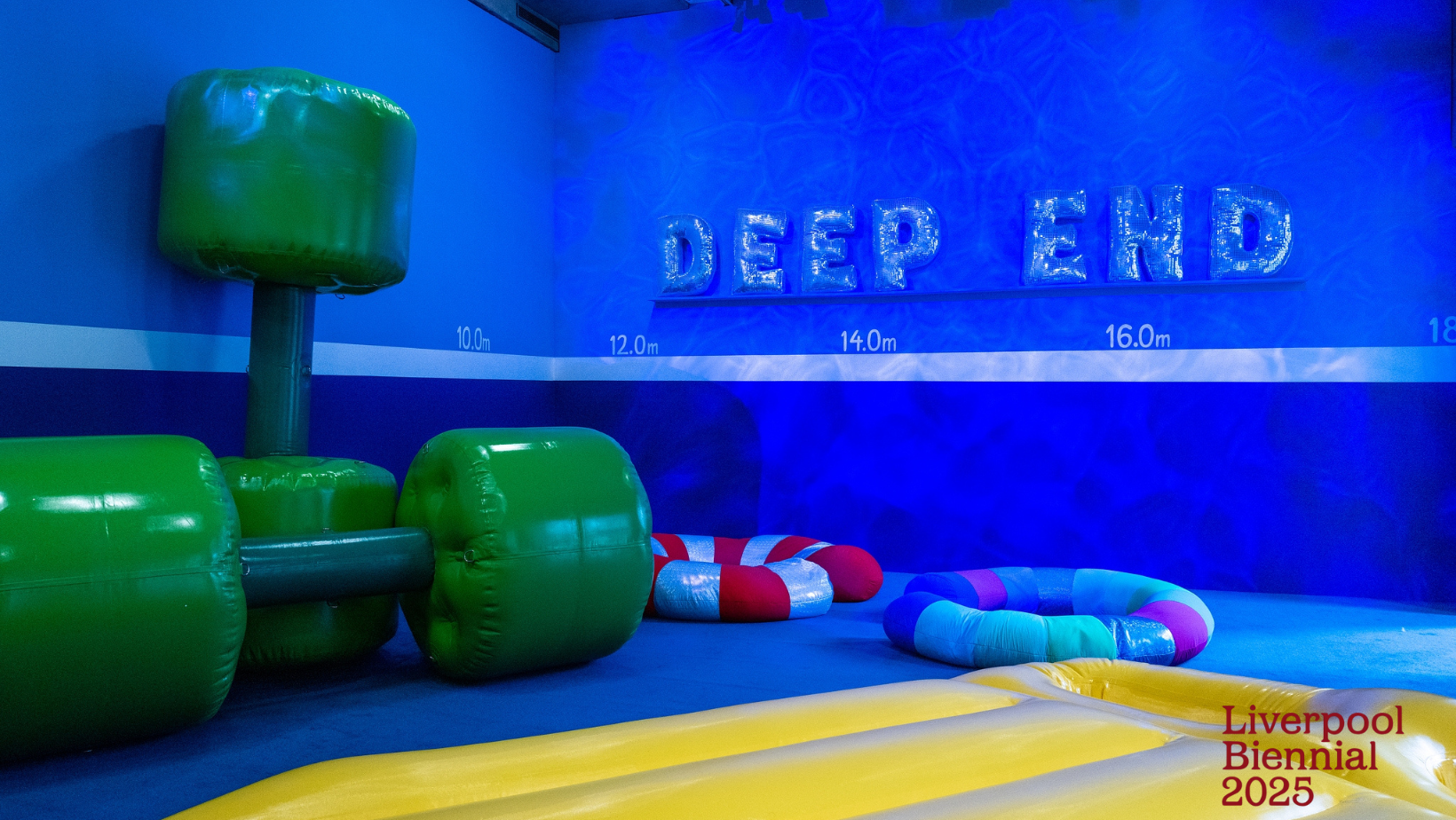 A bright blue wall carries inflated letters reading 'Deep End'. Beneath is a yellow inflatable lilo on the floor next to green inflatable dumbbells