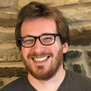 A caucasian person in their twenties with brown hair, a beard and glasses, smiling whilst stood against a brick wall