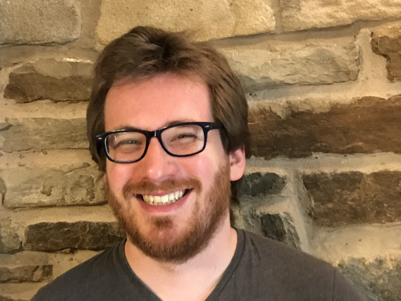 A caucasian person in their twenties with brown hair, a beard and glasses, smiling whilst stood against a brick wall