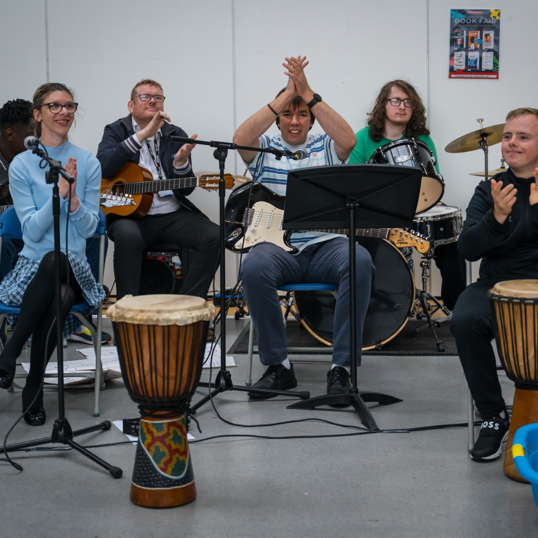 A group of young musicians sit behind music stands. Some have drums, others guitars. They are clapping.