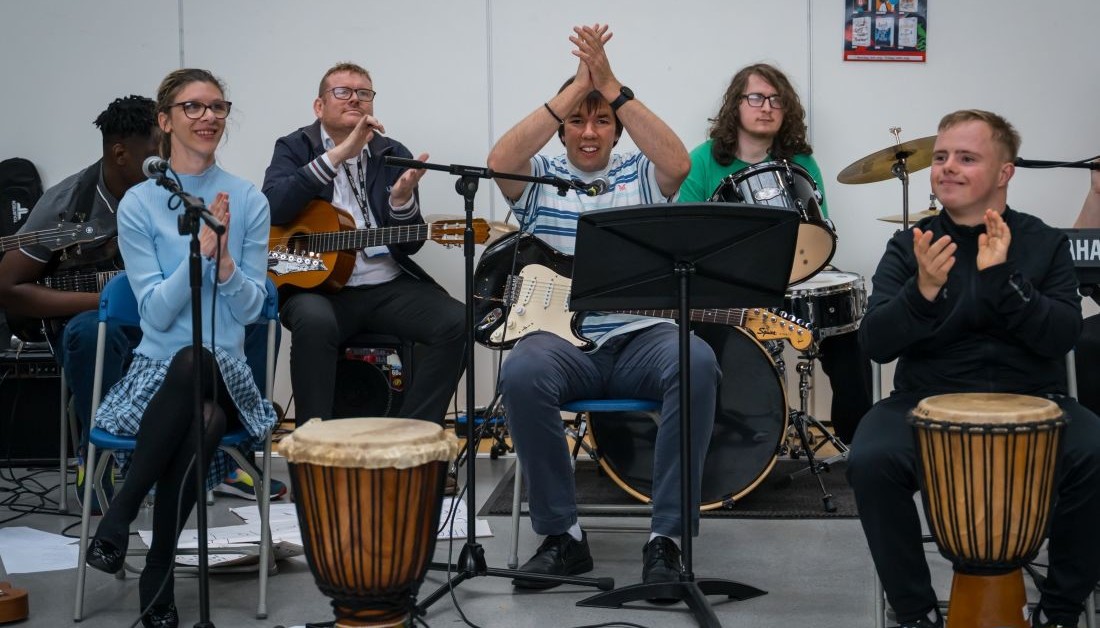 A group of young musicians sit behind music stands. Some have drums, others guitars. They are clapping.
