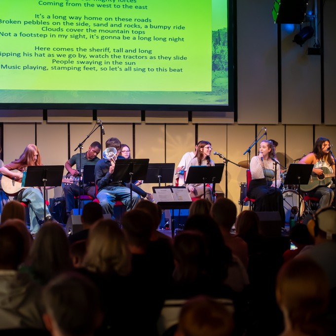 A group of young musicians are gathered on a stage. One is sat at a grand piano. Others have guitars, violin, trumpet and microphones. Behind is a yel