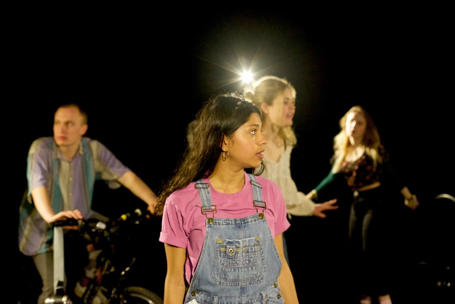 Young people onstage, the foremost of which has long brown hair, wears denim dungarees with a pink tshirt and is looking offstage to her left. 