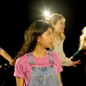 Young people onstage, the foremost of which has long brown hair, wears denim dungarees with a pink tshirt and is looking offstage to her left. 